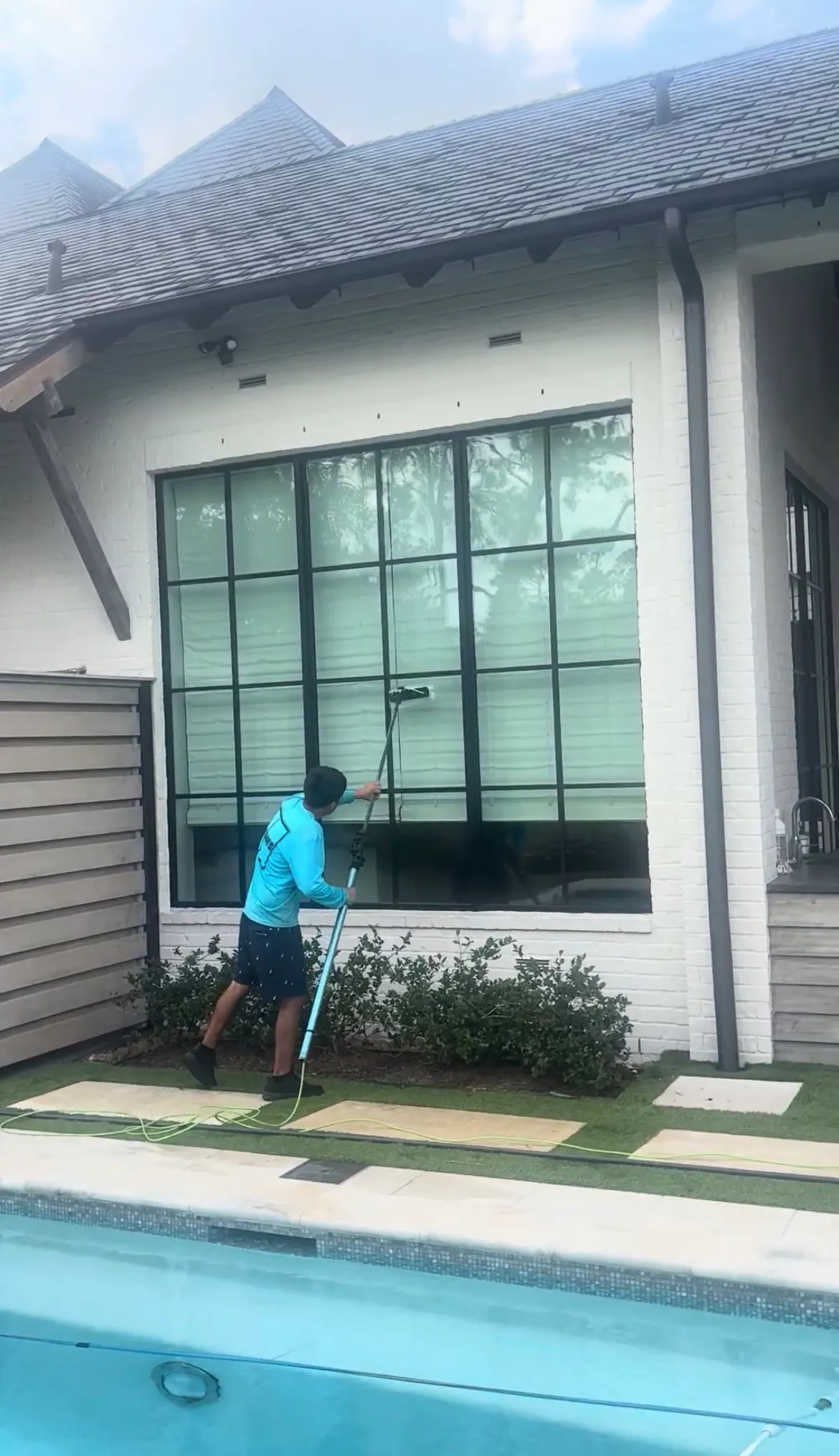 Gleam Clean technician using a water-fed pole to clean tall exterior windows on a white brick home
