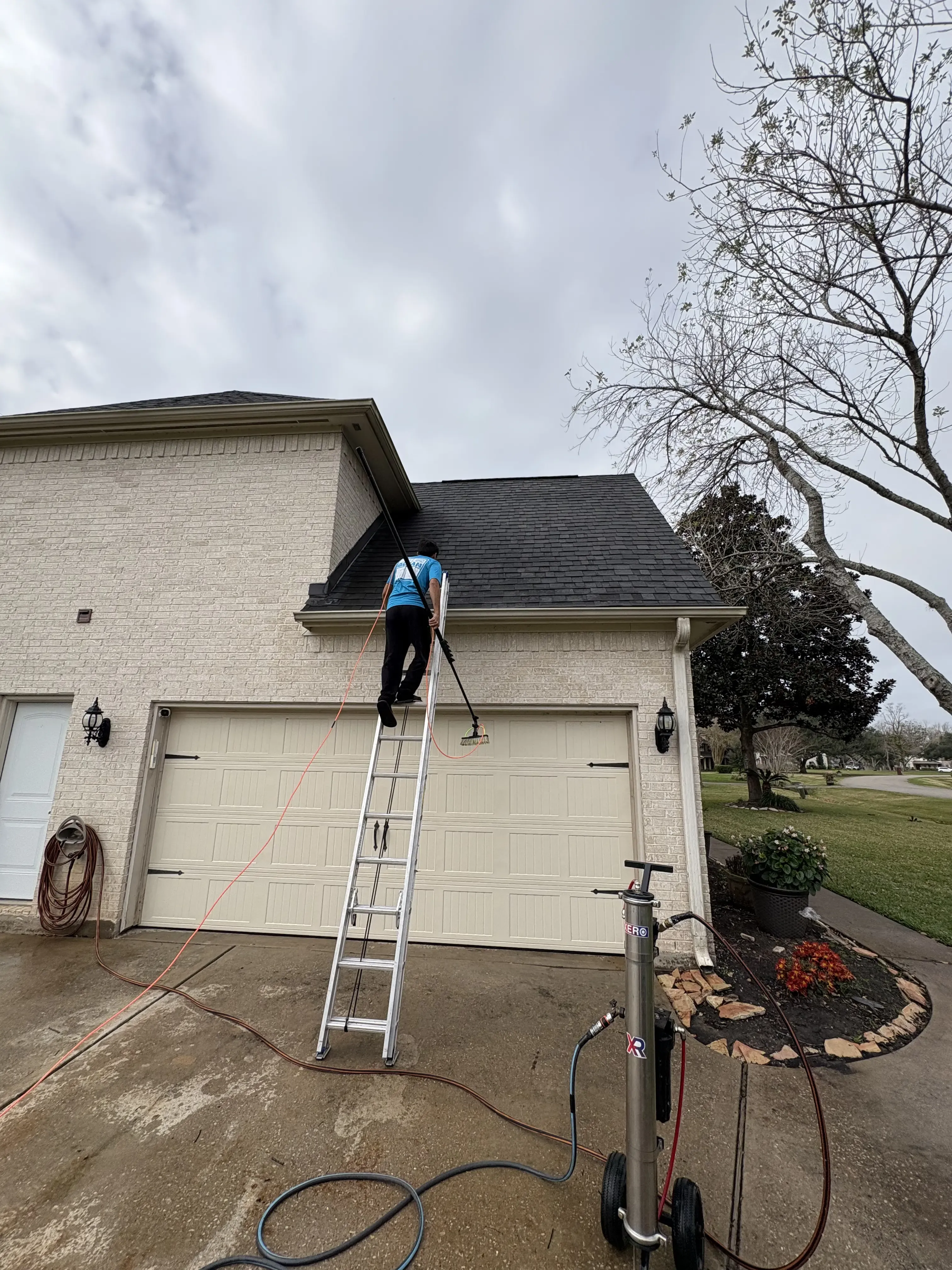 Gleam Clean technician on an extension ladder cleaning the upper windows above the garage of a brick home, with a deionized water tank on the driveway